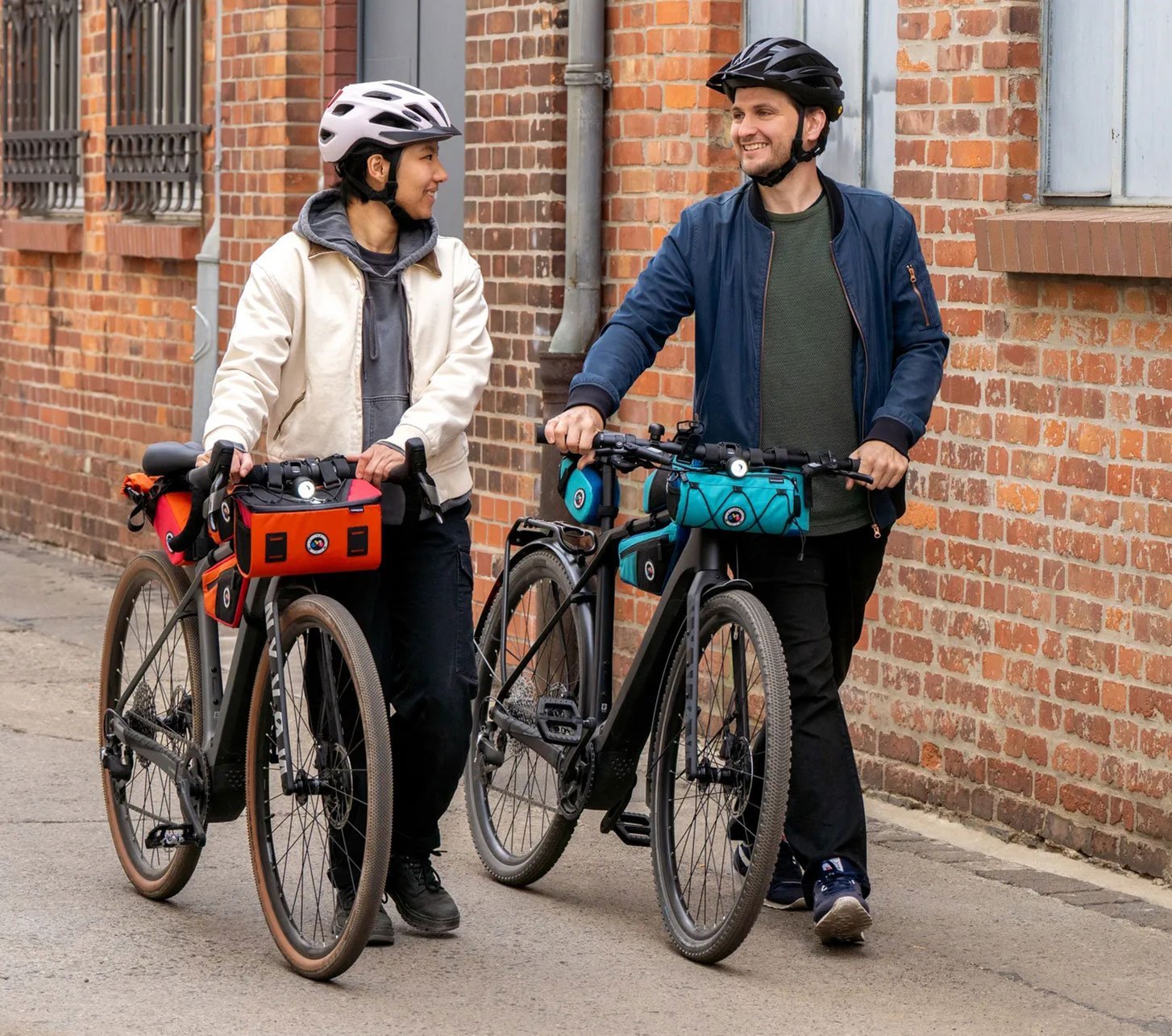 Zwei urbane Radfahrer mit Fahrradhelmen schieben ihre City-Bikes durch eine Backsteinstraße; Lenkertaschen von Contec unterstreichen alltagstaugliches Urban-Cycling und Pendler-Mobilität.