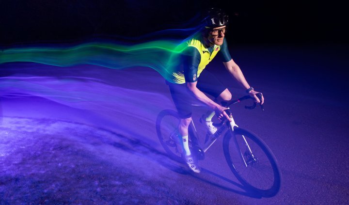 Ein Rennradfahrer mit grellgelber Weste fährt bei Nacht durch einen lilafarbenen Lichtkegel. Er zieht aufgrund der langen Verschlusszeit eine Spur bunter Lichter hinter sich her. 