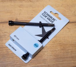 Close-up: A package containing two Schwalbe Clik Valve tubeless valves lying on a brown wooden surface.