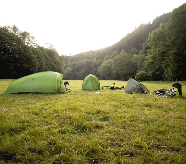 3 VAUDE tents pitched on a meadow surrounded by trees. Next to each tent is a bicycle.