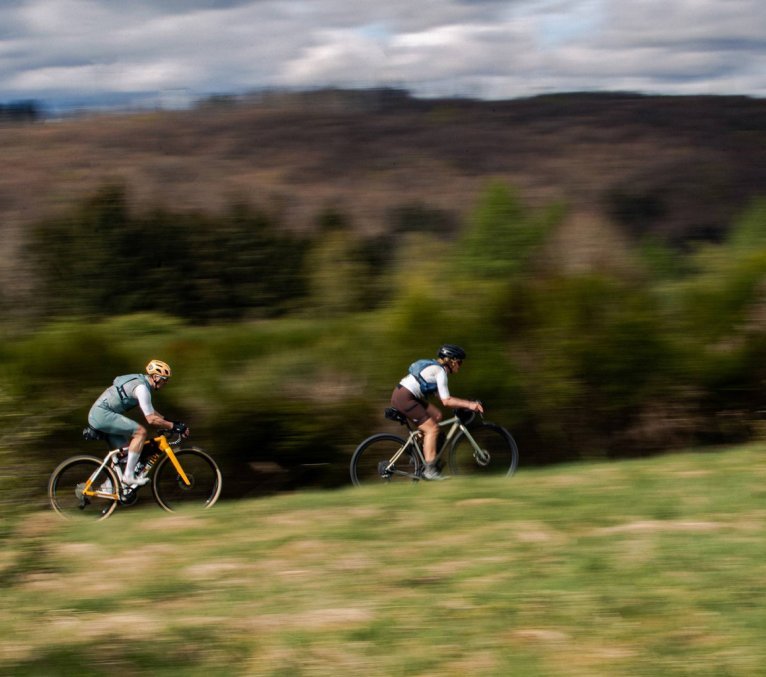 Deux cyclistes gravel roulent rapidement à travers une prairie vallonnée, arrière-plan flou soulignant le mouvement.