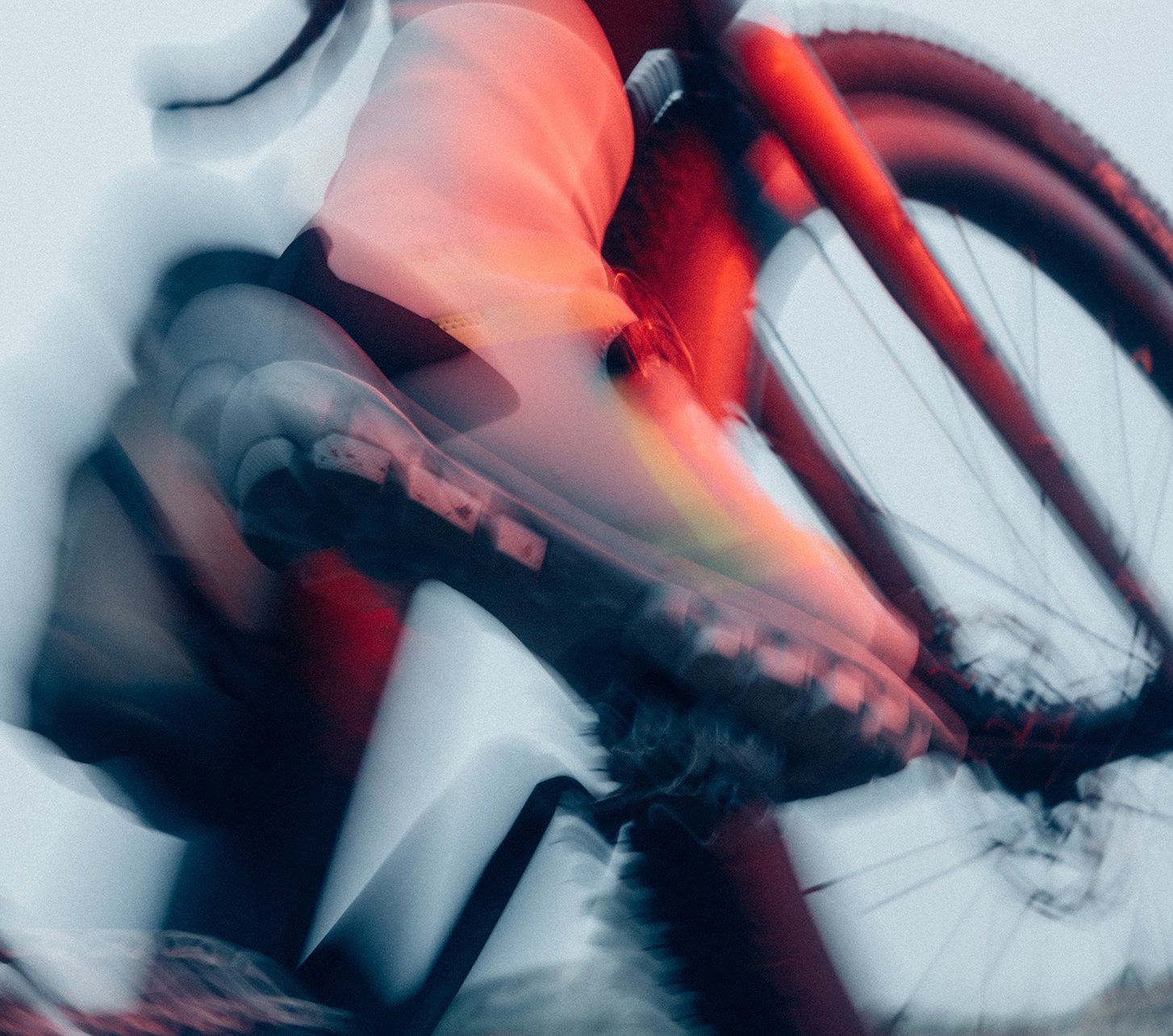 Dynamische Nahaufnahme eines Fahrradschuhs auf dem Pedal mit deutlicher Bewegungsunschärfe. Fahrradrahmen und Reifen sind im Hintergrund erkennbar, während rote und blaue Lichtreflexe die Bewegung betonen.