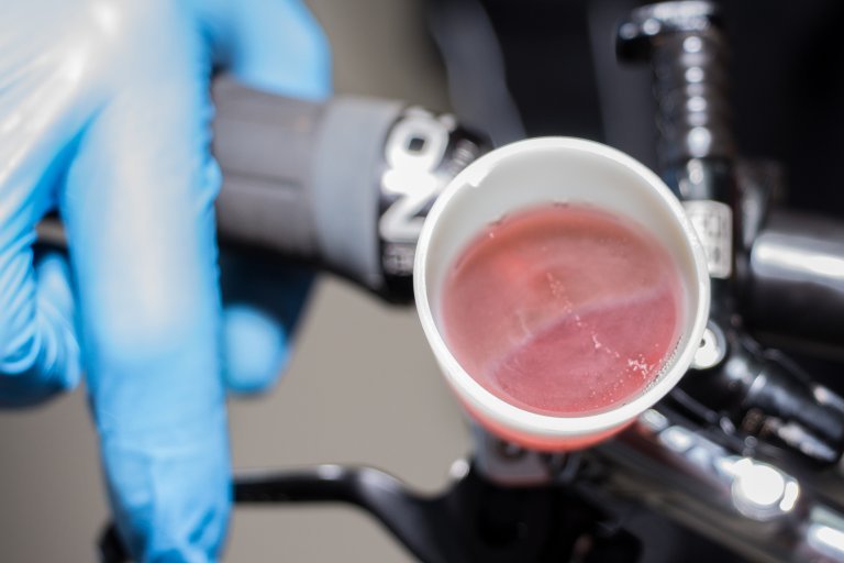 A small white funnel attached to the brake lever of a bicycle contains a pink fluid with air bubbles rising in it.