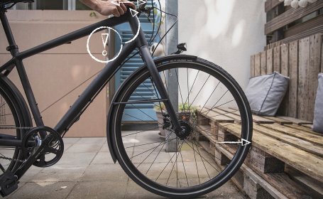 Die Hand umgreift den Steuersatz am Kontaktpunkt zum Rahmen und schiebt das Fahrrad gleichzeitig mit leichtem Druck gegen den festen Gegenstand.