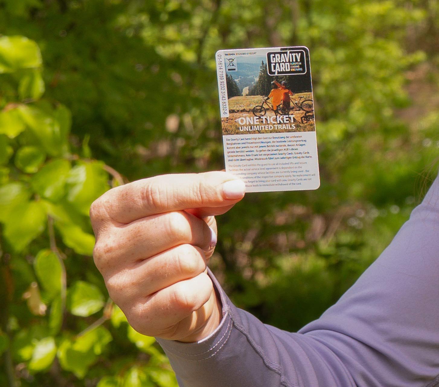 Person hält eine Gravity Card 2026 im Wald und zeigt auf das Ticket für Bikepark- und Trail-Zugang, Gewinnspielmotiv im Outdoor-Setting.