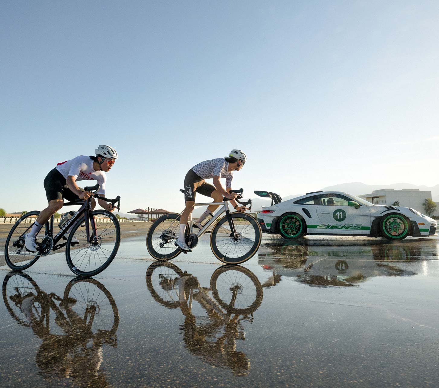 Dos ciclistas de carretera montando bicicletas Cannondale CAAD14 en una pista mojada junto a un coche deportivo Porsche 911 GT3 RS.