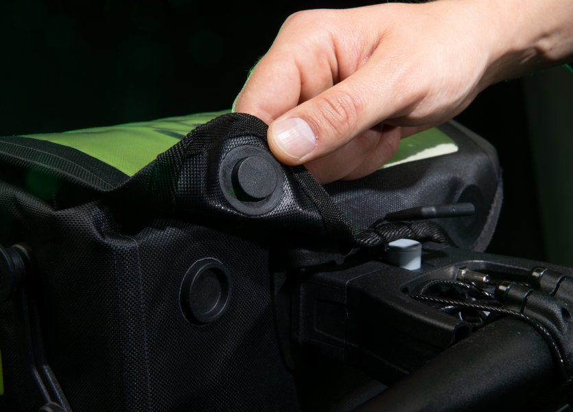 Eine Hand schließt eine schwarze Fahrradtasche mit grünen Akzenten. Die Tasche hat ein beleuchtetes, perforiertes Design. Der Hintergrund ist dunkel.