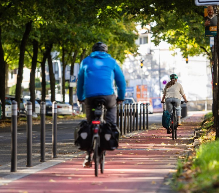 Deux cyclistes roulent sur une piste cyclable rouge bordée d’arbres ; des bornes séparent la piste de la chaussée.