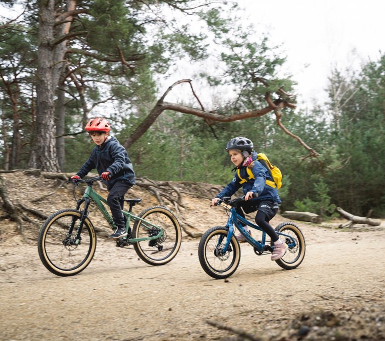 Three children ride on SUPURB and Specialized kids mountain bikes along a forest path.