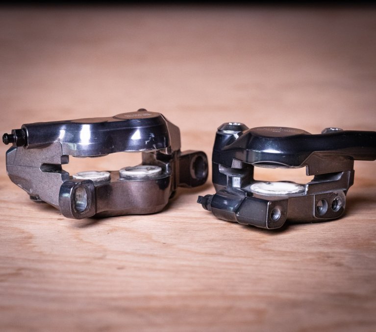 Dos pinzas de freno para frenos de disco de bicicleta están colocadas una al lado de la otra sobre una tabla de madera de tono cálido.