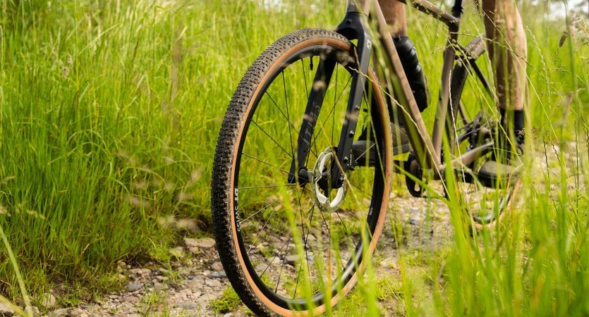 Roue avant d’un vélo gravel sur chemin caillouteux, pneu à crampons marqués et flanc beige en action dans un environnement verdoyant.