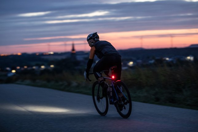 Un ciclista en una bicicleta de carretera pedalea en una vía al anochecer, con luces alimentadas por batería en el manillar y el poste del sillín, mientras las luces de una ciudad brillan al fondo.