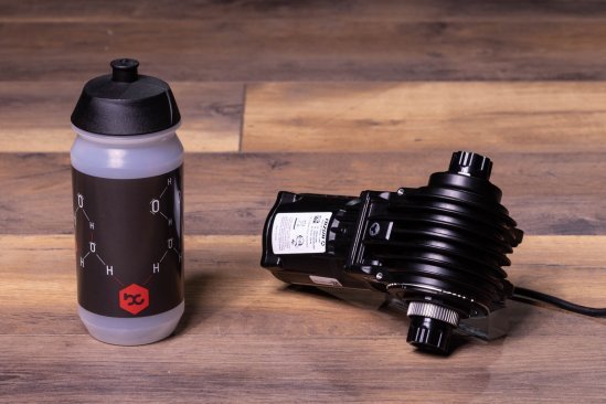 A Fazua Ride 60 e-bike drive lies on a dark wooden floor. A bike water bottle stands next to it
