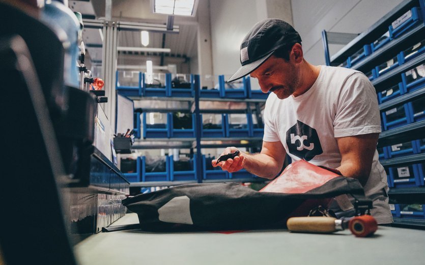 Andi à l'un des postes de travail de l'atelier chez ORTLIEB. Il tient un fermoir de sacoche dans la main. Devant lui se trouve une sacoche à bagages.