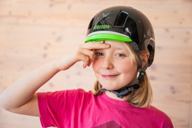 A blonde girl is wearing a black bike helmet from the brand Affenzahn. She places two fingers over her eyebrows to show where the helmet should sit.