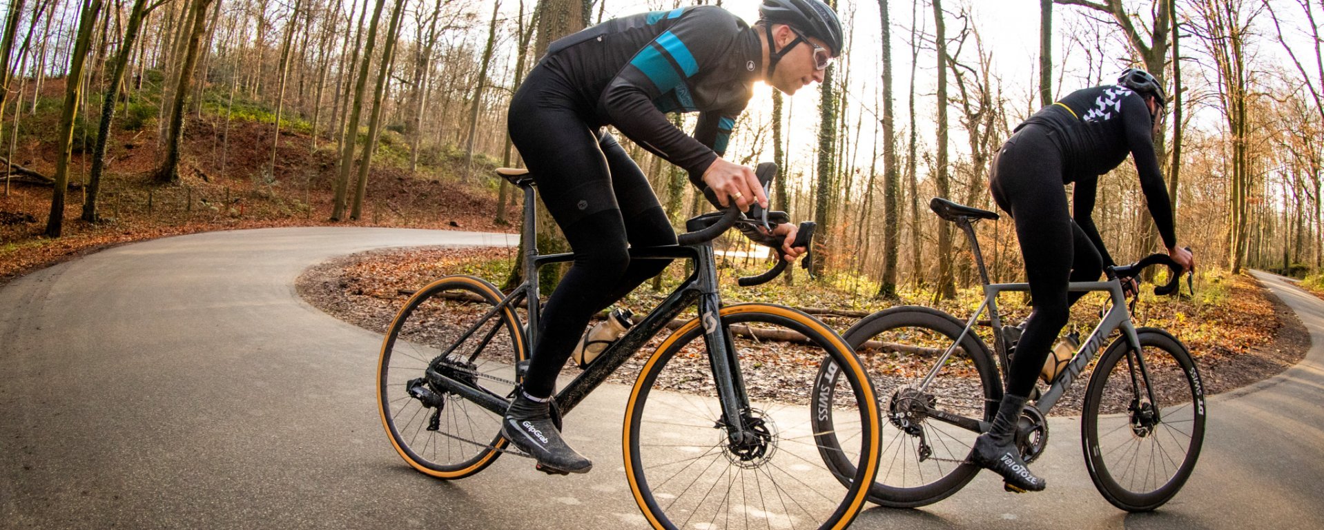 Two cyclists in black sportswear with helmets are riding on a winding road through a forest with autumn-colored trees. The bikes have deep rims and brown tires.