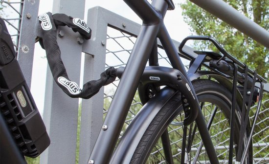 Bicicleta con candado de cuadro bloqueando la rueda trasera y una cadena adicional asegurada a una valla metálica.