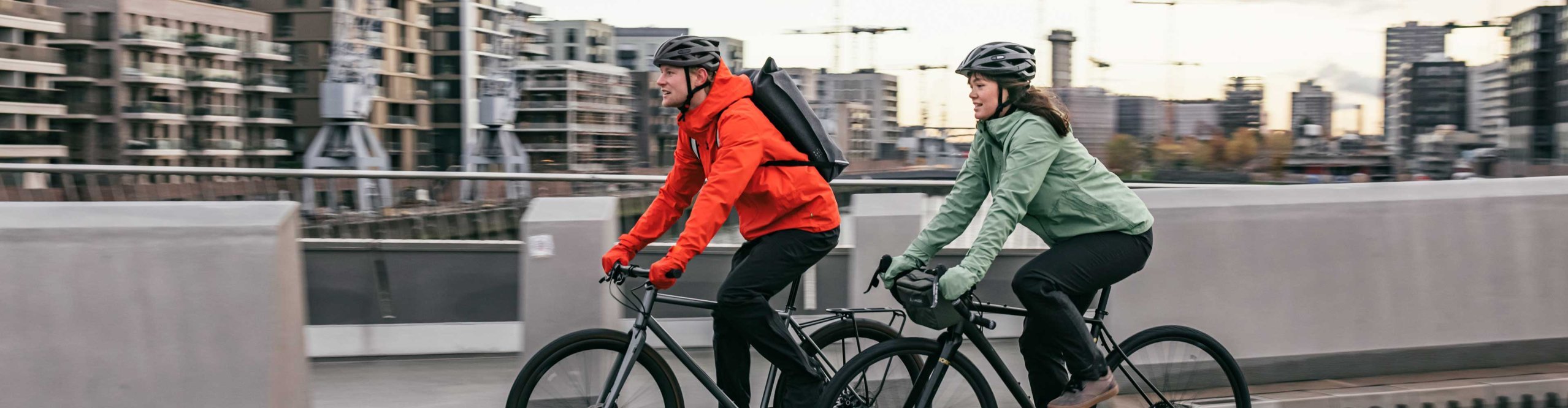 Two cyclists wearing Vaude rain jackets ride their bikes through Hamburg’s HafenCity district.