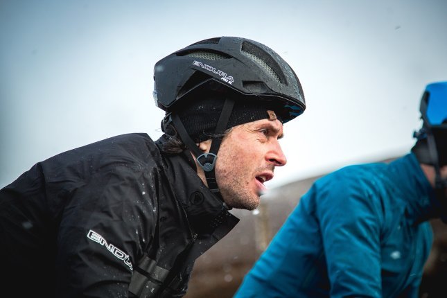 A man wearing a black cycling jacket and a black bike helmet is riding in rainy weather, with another cyclist in the background.