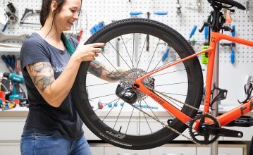 Isa from the bc Service Department mounts the rear Zipp 303 wheel on an orange Factor LS gravel bike. The bicycle hangs on a repair stand.