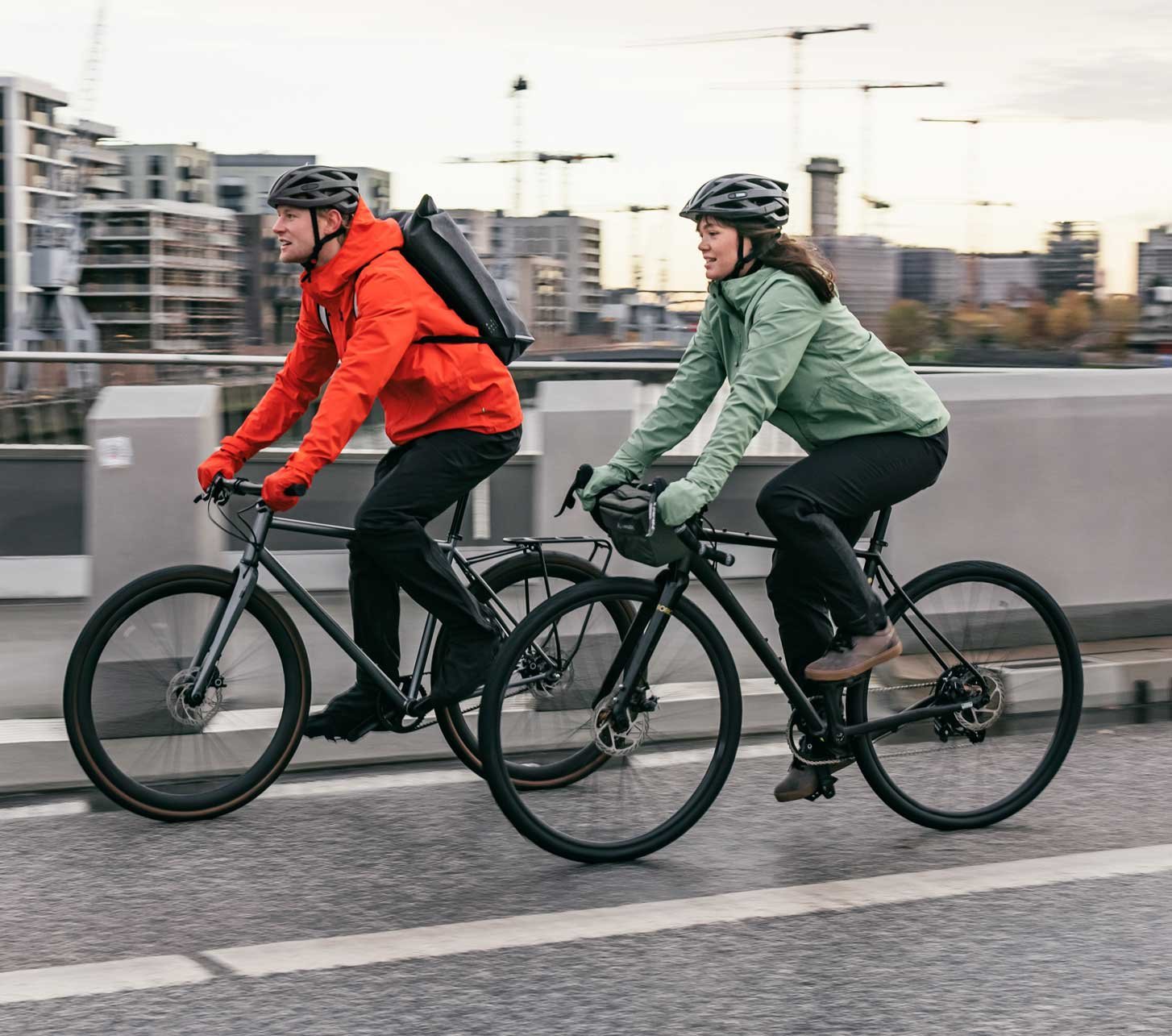 Two cyclists wearing Vaude rain jackets ride their bikes through Hamburg’s HafenCity district.