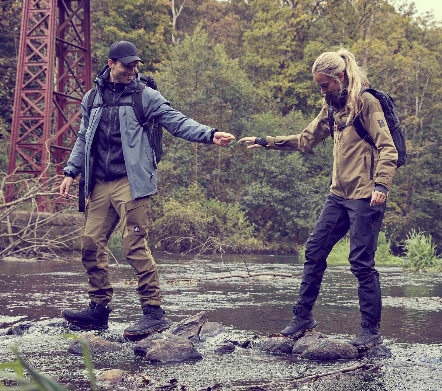 Two hikers wearing outdoor clothing and backpacks cross a stream beneath an iron bridge