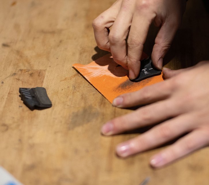 A hand roughens a brake pad with sandpaper on a workbench.