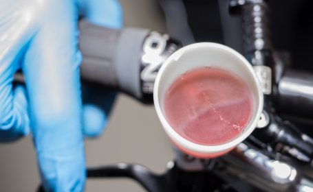 Ein kleiner weißer Trichter, der am Bremshebel eines Fahrrads befestigt ist, enthält eine rosa Flüssigkeit, in der Luftblasen aufsteigen.