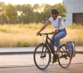 Femme portant un casque roulant à vélo urbain sur une route calme à la lumière du soir ; sacoche fixée au porte-bagages, prairie et arbres en arrière-plan.