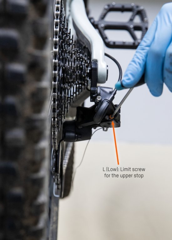 The rear derailleur can be seen from behind. A hex wrench is put on the outer stop. An orange arrow marks the L-screw. 