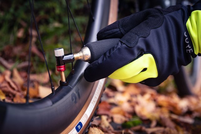 A gloved hand screws a CO₂ cartridge into a pump head attached to a bicycle wheel. Autumn leaves blur softly in the background.