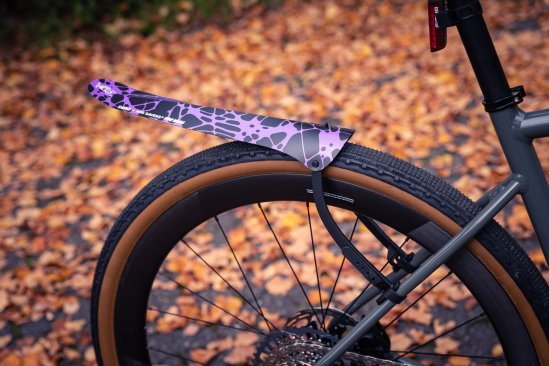 A black Ass Savers "Win Wing" mudguard with a purple splatter design mounted on the rear wheel of a grey bicycle.