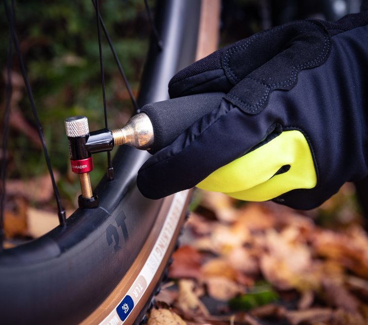 A gloved hand screws a CO₂ cartridge into a pump head attached to a bicycle wheel. Autumn leaves blur softly in the background.