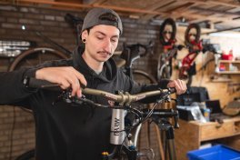 Eine Person in einer Werkstatt überprüft die Bremsleitungen eines Fahrrads, das an einem Ständer befestigt ist, mit Werkzeugen und weiteren Fahrrädern im Hintergrund.