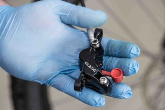 A hand wearing blue gloves holds a brake caliper of a hydraulic disc brake with a red bleed screw, ready for maintenance, with a bicycle in the background.