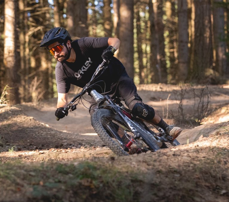 A mountain biker rides on a forest trail, surrounded by trees and a dirt path.