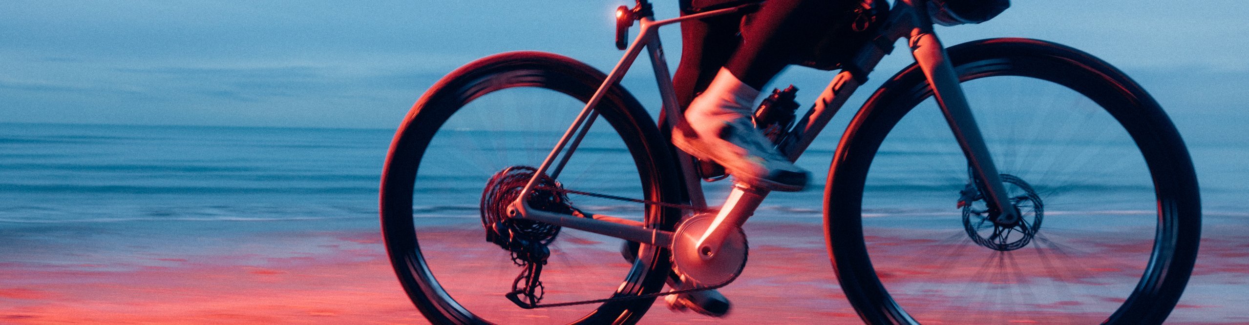 Nahaufnahme eines fahrenden Fahrrads am Strand bei Dämmerlicht. Die Aufnahme zeigt die rot beleuchteten Laufräder und die Beine der radelnden Person, während das Meer im Hintergrund ruhig erscheint.