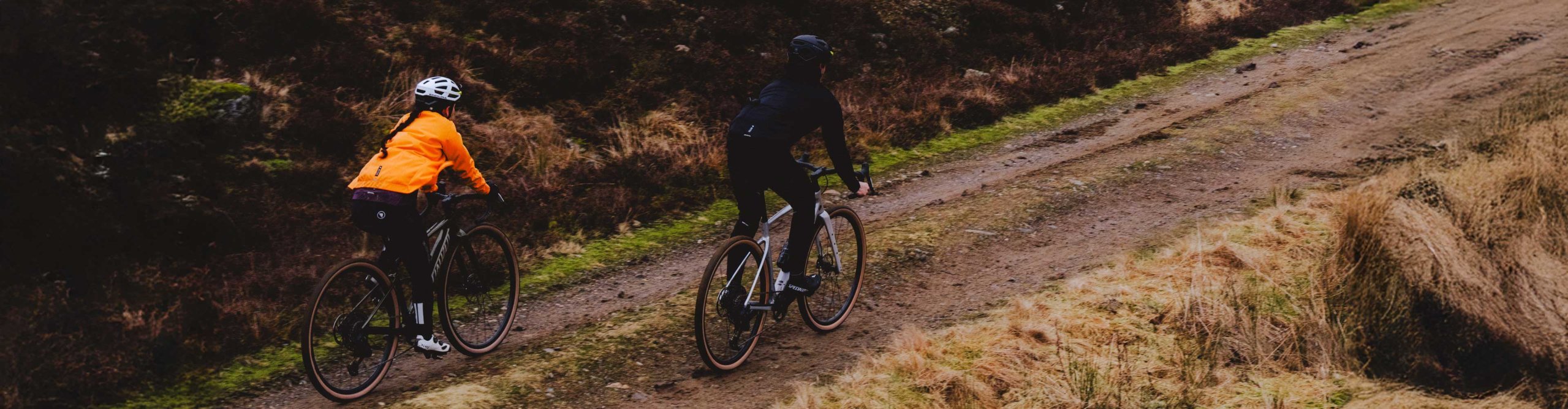 Deux cyclistes gravel portant des vestes Endura roulent sur un chemin humide à travers un paysage vallonné.