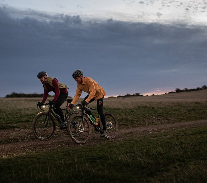 Dos ciclistas con luces en sus bicicletas pedalean por un camino de tierra al atardecer, rodeados de campos y un cielo nublado.