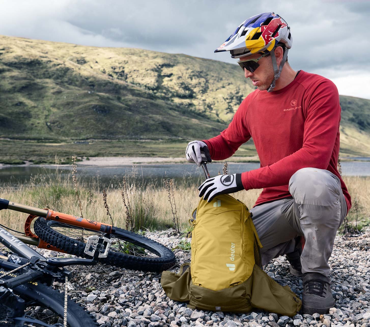 Mountainbiker kniet auf Schotter neben seinem MTB und öffnet einen gelben Deuter Rucksack in alpiner Landschaft