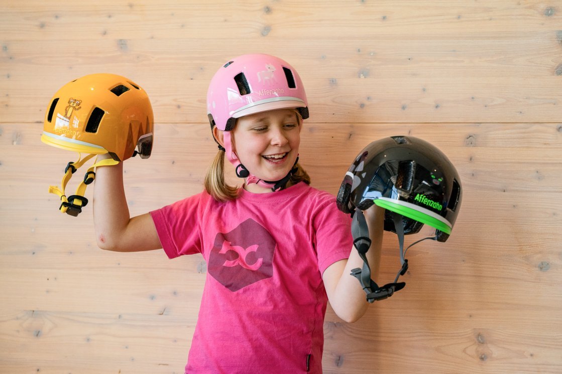 A blonde, about 10-year-old girl in a pink BC t-shirt stands in front of a light wooden wall, wearing a pink bike helmet and holding another helmet in each hand.