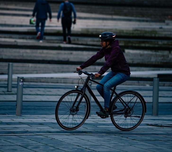 Un ciclista con luces en su bicicleta y casco pedalea en una plaza pavimentada de la ciudad por la noche, mientras dos personas caminan al fondo.