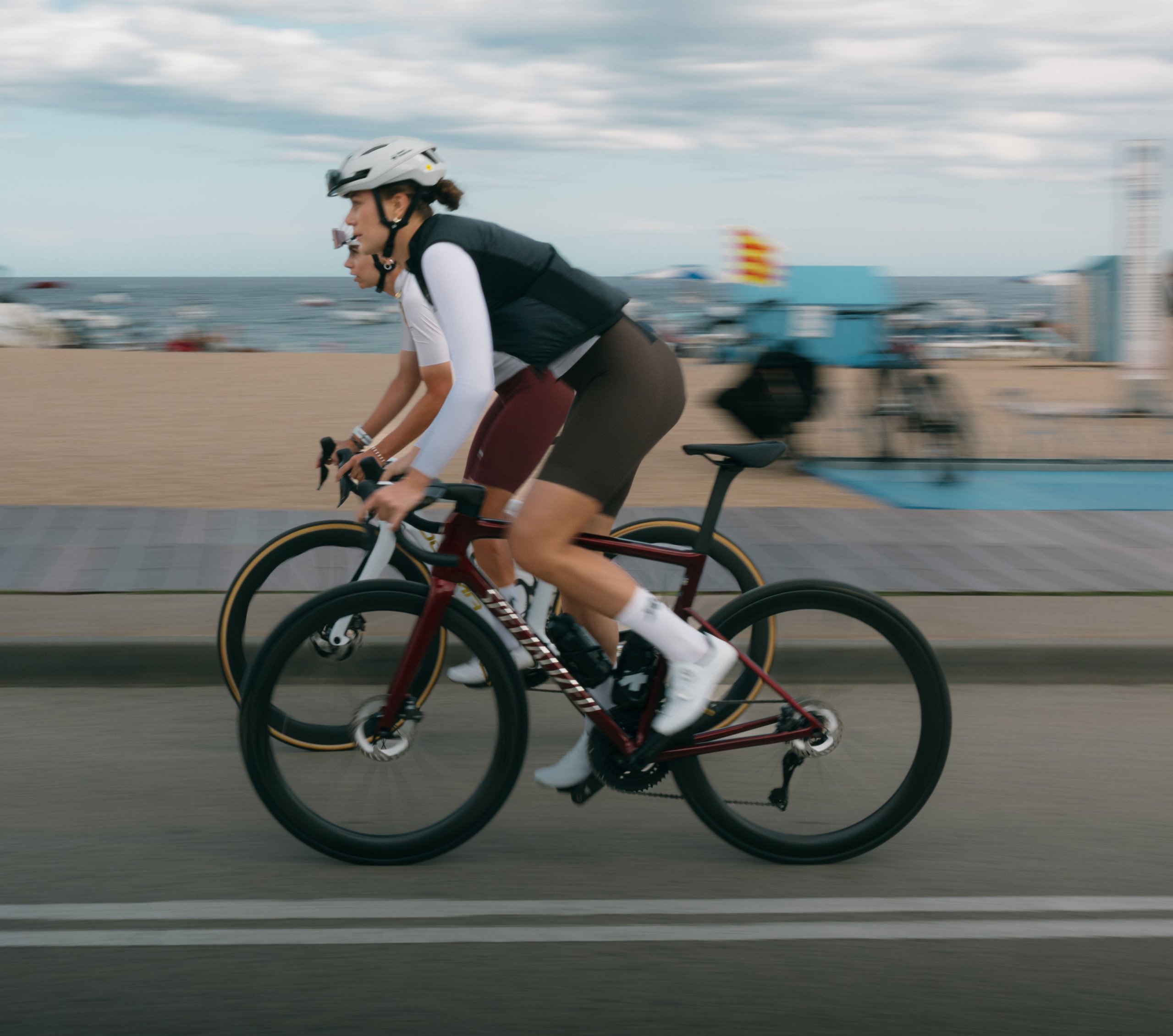 Dos ciclistas de carretera con outfit de Assos ruedan por un paseo marítimo, con playa y mar de fondo.