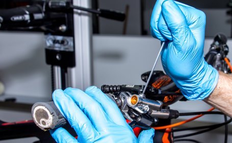 Eine Hand mit blauen Handschuhen dreht die Entlüftungsschraube an einem Bremshebel eines Fahrrads heraus, während das Fahrrad an einem Ständer befestigt ist.