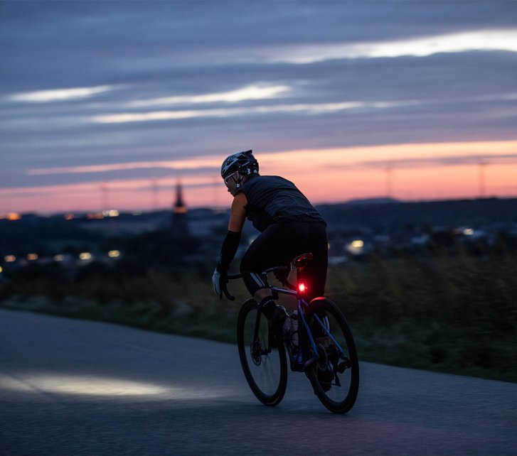 Un ciclista en una bicicleta de carretera pedalea en una vía al anochecer, con luces alimentadas por batería en el manillar y el poste del sillín, mientras las luces de una ciudad brillan al fondo.
