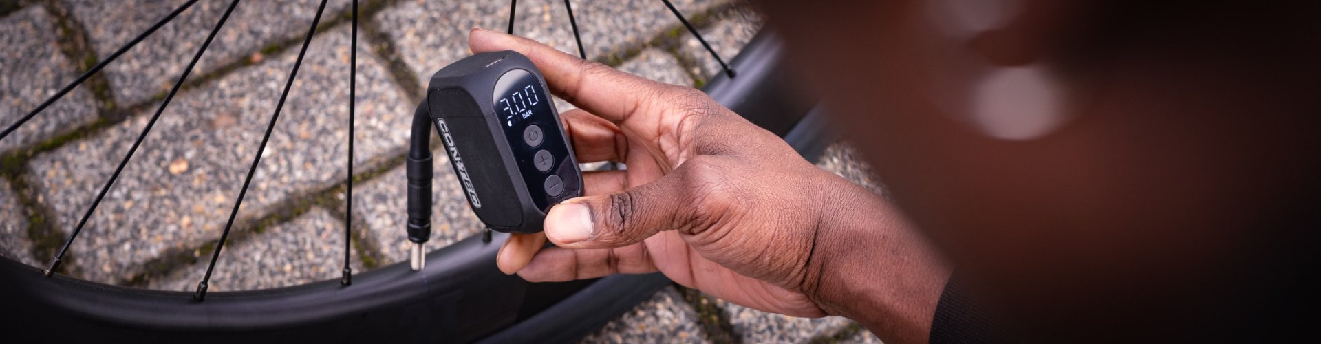 Eine Männerhand mit dunkler Haut hält einen Mini-Kompressor, der am Ventil eines Rennrad-Laufrads angeschlossen ist. Das Rad liegt auf Steinboden