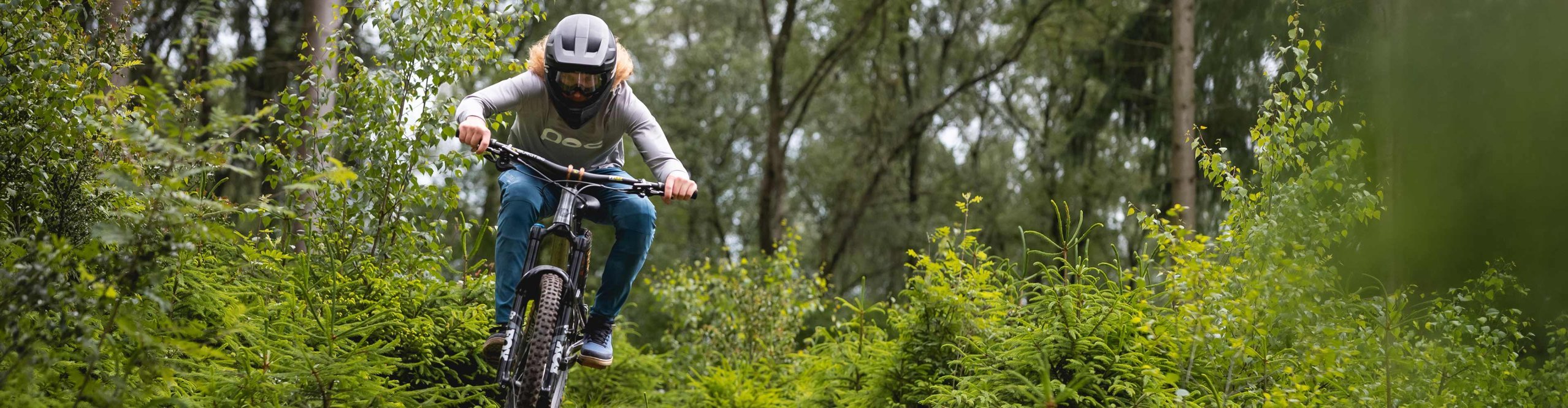 Vététiste roulant sur un sentier forestier avec racines et petites marches – scène illustrant des vêtements VTT respirants pour le printemps et l’été.
