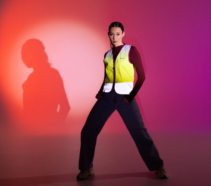 Eine junge Frau posiert vor einem bunt orange und pink angestrahlten Papierhintergrund. Zu einer schwarzen Hose trägt sie eine Leuchtweste von Wowow