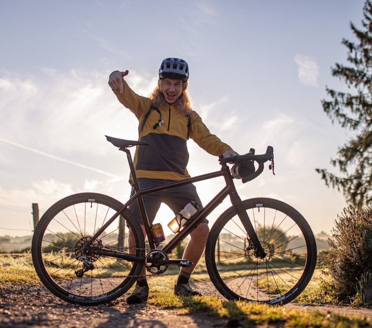 Gegenlichtaufnahme auf einem geschotterten Wirtschaftsweg: bc Mitarbeiter Kai steht stolz hinter seinem neuen Gravel-Bike und zeigt mit dem Finger darauf.