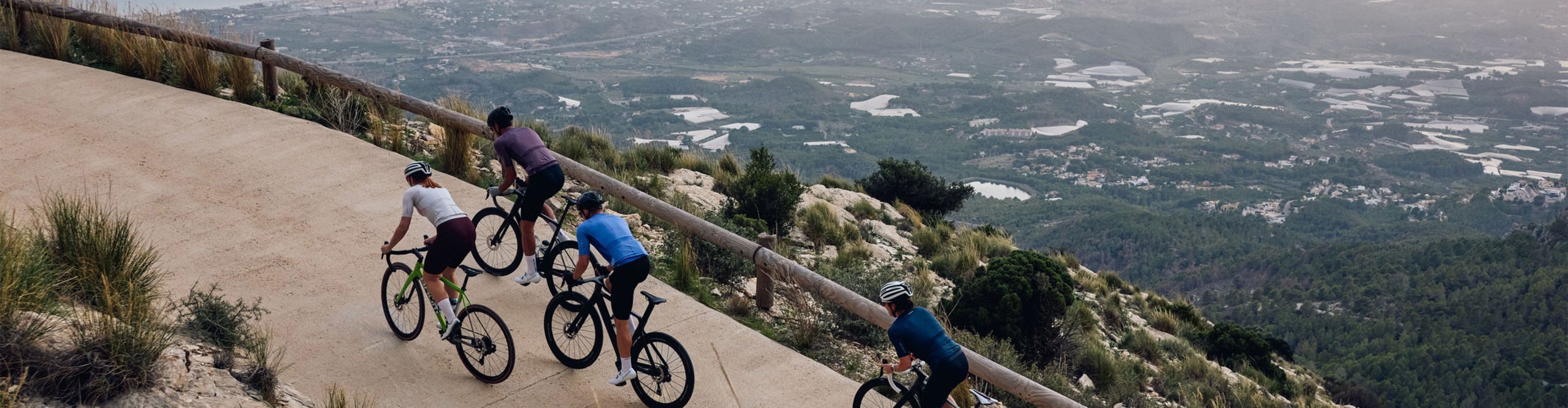 Eine 4 köpfige Gruppe RennradfahrerInnen fahren im Wiegetritt eine asphaltierte Straße hoch. Im Hintergrund ist eine am Meer gelegene Stadt und Berge zu sehen.